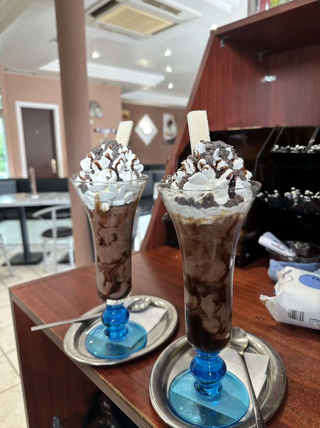 Zwei Schokoladen-Eisbecher mit Sahne und Waffel in Gläsern mit blauem Fuß auf der Theke, dahinter der Gastraum